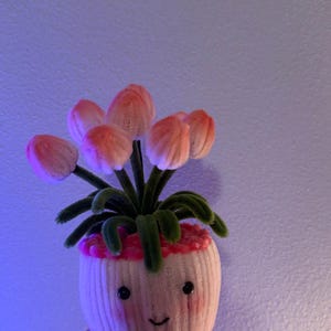 May include: A whimsical, handmade felt flower pot with a smiling face. The pot holds several pink and orange felt flowers with green stems and leaves. The pot is held in a hand.