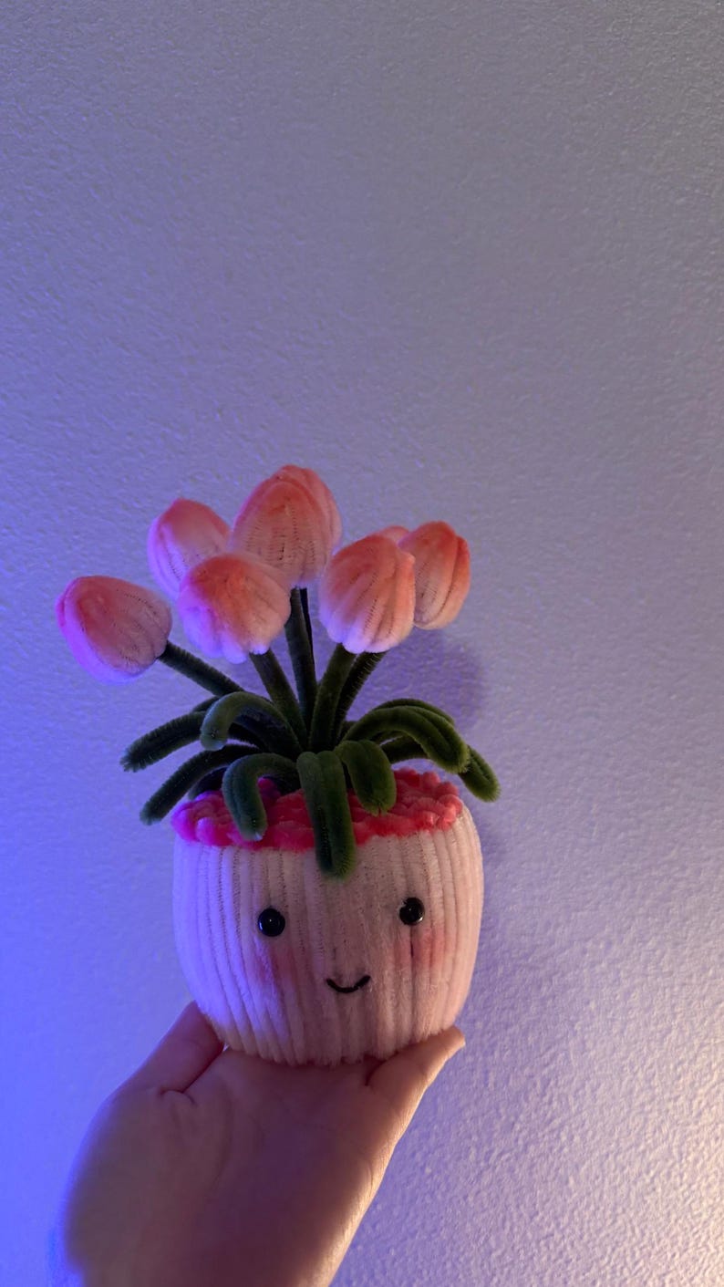 May include: A whimsical, handmade felt flower pot with a smiling face. The pot holds several pink and orange felt flowers with green stems and leaves. The pot is held in a hand.