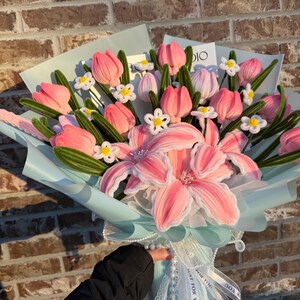 May include: A bouquet of pink and white fabric flowers, including tulips and lilies, wrapped in light blue paper and tied with white ribbons. The bouquet is held by a person wearing a black jacket. The background is a brick wall.
