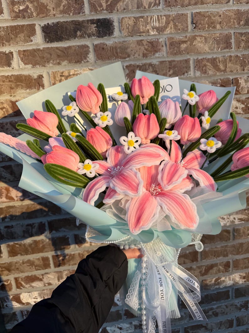 May include: A bouquet of pink and white fabric flowers, including tulips and lilies, wrapped in light blue paper and tied with white ribbons. The bouquet is held by a person wearing a black jacket. The background is a brick wall.