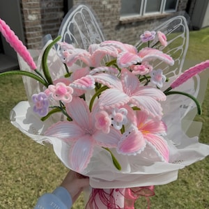 May include: A bouquet of pink and white handmade flowers, including lilies and smaller blossoms, wrapped in white paper and adorned with pink ribbons. The flowers are made of a soft, textured material.