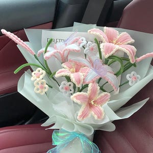 May include: A bouquet of handmade pink and white crochet lilies and small flowers, wrapped in light blue paper and tied with a blue iridescent ribbon. The bouquet includes a butterfly and the word "PRETTY" on a card.