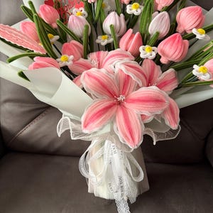 May include: A bouquet of pink and white fabric flowers, including tulips and larger blossoms, wrapped in light blue paper. Green stems and small white flowers accent the arrangement. White ribbon with text is tied around the base.