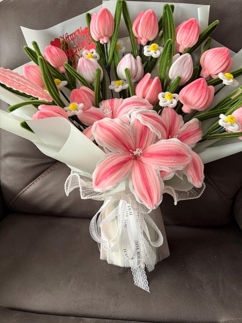 May include: A bouquet of pink and white fabric flowers, including tulips and larger blossoms, wrapped in light blue paper. Green stems and small white flowers accent the arrangement. White ribbon with text is tied around the base.
