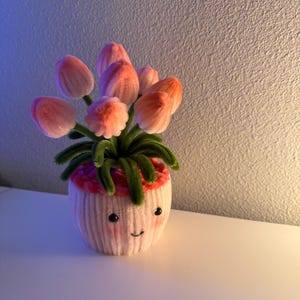 May include: A handmade, whimsical flower pot with a smiling face, holding a bouquet of pink and orange tulips. The pot is light pink with a red rim and green stems. The flowers are made of soft, textured material.
