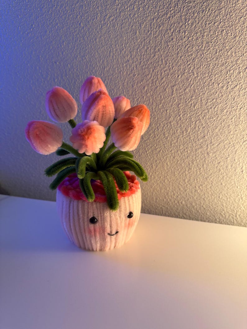 May include: A handmade, whimsical flower pot with a smiling face, holding a bouquet of pink and orange tulips. The pot is light pink with a red rim and green stems. The flowers are made of soft, textured material.