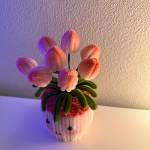 May include: A handmade floral arrangement featuring pink tulip-like flowers with green stems and leaves, arranged in a white cupcake-shaped vase. The vase has a smiling face design. The arrangement is made of soft, fuzzy material.