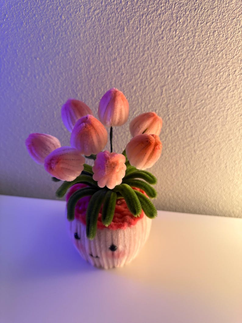 May include: A handmade floral arrangement featuring pink tulip-like flowers with green stems and leaves, arranged in a white cupcake-shaped vase. The vase has a smiling face design. The arrangement is made of soft, fuzzy material.