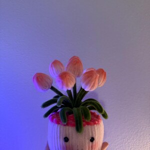 May include: A whimsical, handmade flower pot with a smiling face. The pot is white with pink accents and holds several pink and green fabric tulips. The pot is held in a hand against a blue background.
