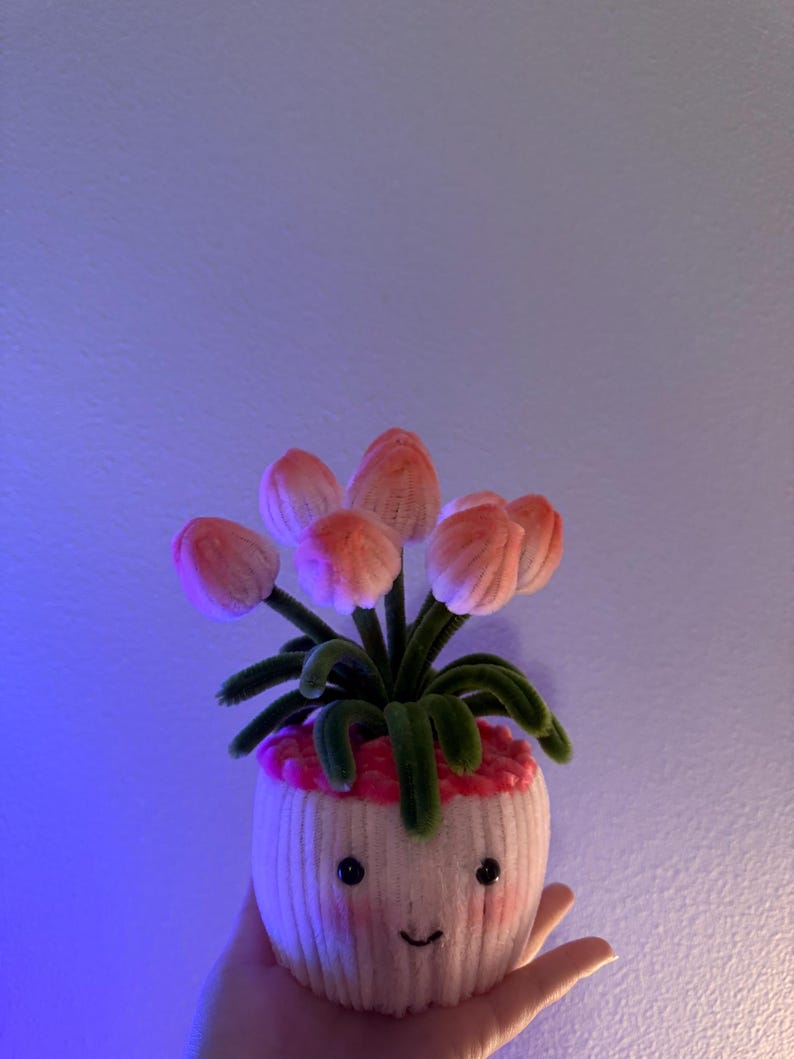 May include: A whimsical, handmade flower pot with a smiling face. The pot is white with pink accents and holds several pink and green fabric tulips. The pot is held in a hand against a blue background.