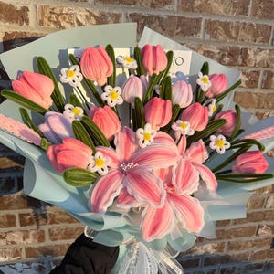 May include: A bouquet of pink and white fabric flowers, including tulips and lilies, wrapped in light blue paper. The bouquet is tied with a white ribbon and lace, creating a delicate and handcrafted floral arrangement.
