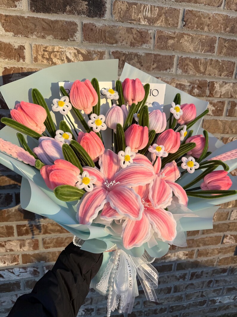 May include: A bouquet of pink and white fabric flowers, including tulips and lilies, wrapped in light blue paper. The bouquet is tied with a white ribbon and lace, creating a delicate and handcrafted floral arrangement.