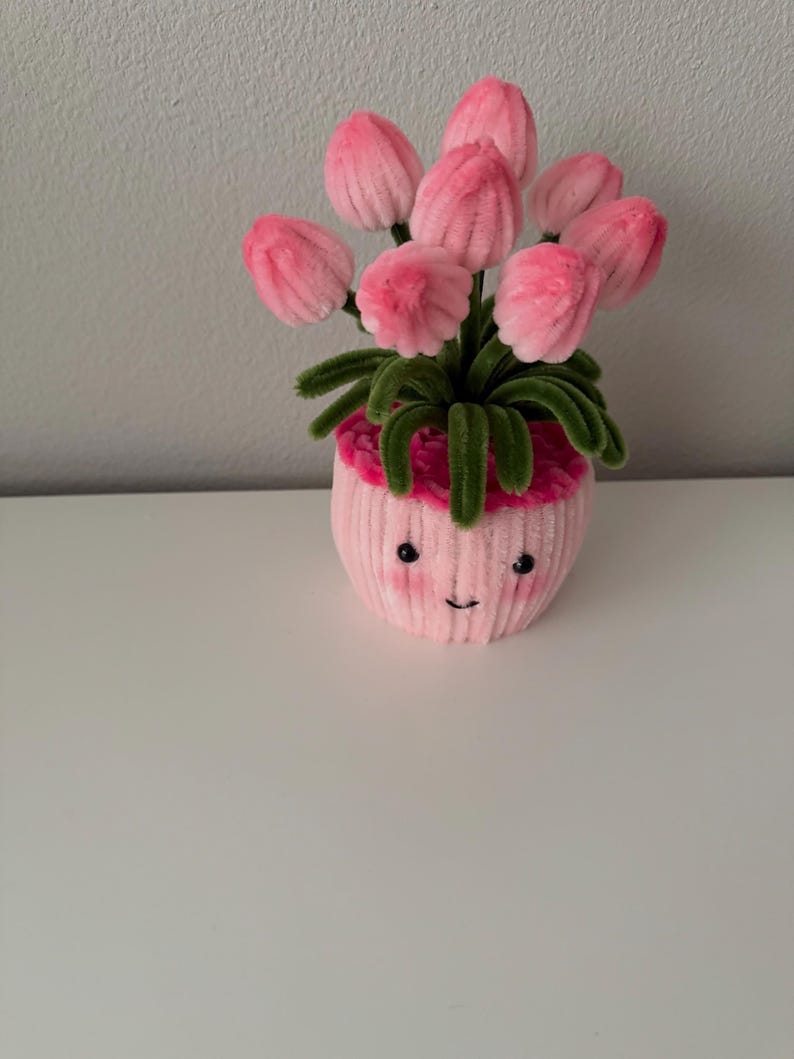 May include: A pink, plush flower pot with a smiling face, holding a bouquet of pink tulips with green stems and leaves. The pot has a ribbed texture and a pink trim around the top.