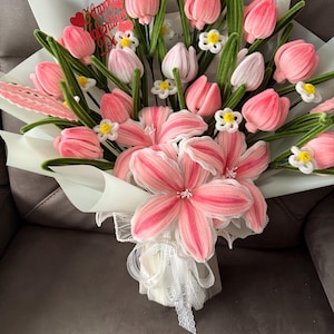 May include: A bouquet of pink and white tulips and lilies, with green stems and small white flowers. The bouquet is wrapped in white paper and tied with a white ribbon. A red heart-shaped decoration with the words "Happy Valentine's Day" is included.