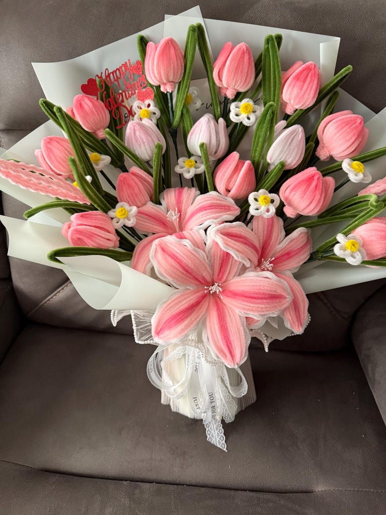 May include: A bouquet of pink and white tulips and lilies, with green stems and small white flowers. The bouquet is wrapped in white paper and tied with a white ribbon. A red heart-shaped decoration with the words "Happy Valentine's Day" is included.