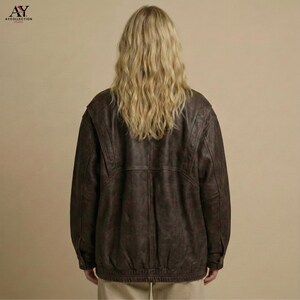 Puede incluir: Chaqueta de cuero marr&oacute;n con dise&ntilde;o tipo bomber. La chaqueta tiene dobladillo y pu&ntilde;os acanalados, y cremallera frontal. La modelo lleva la chaqueta con pantalones beiges. El logo "AY COLLECTION" est&aacute; en la esquina superior izquierda.