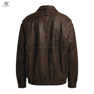 Puede incluir: Chaqueta de cuero marr&oacute;n con dise&ntilde;o tipo bomber. La chaqueta presenta un cuello, una peque&ntilde;a trabilla en la parte trasera del cuello y un dobladillo acanalado. La parte trasera de la chaqueta es visible, mostrando las costuras y los detalles de los paneles.