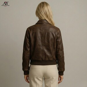 May include: A brown leather bomber jacket with a ribbed hem and cuffs. The jacket has a classic collar and a textured appearance. The model is wearing beige pants. The jacket is a stylish outerwear option.