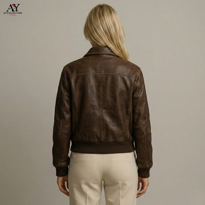 May include: A brown leather bomber jacket with a ribbed hem and cuffs. The jacket has a classic collar and a textured appearance. The model is wearing beige pants. The jacket is a stylish outerwear option.