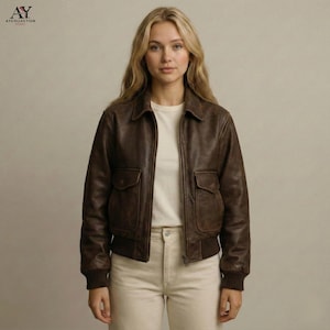 May include: A brown leather bomber jacket with two front pockets, a collar, and a ribbed hem and cuffs. The jacket is zipped up and worn over a white t-shirt and light-colored pants. The jacket has a vintage look.