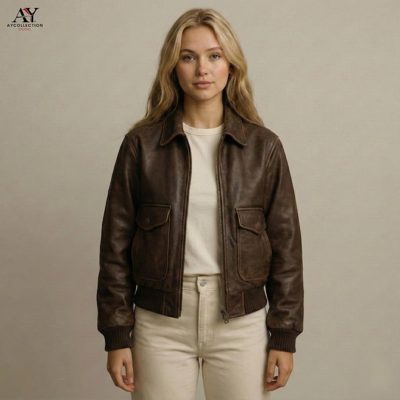 May include: A brown leather bomber jacket with two front pockets, a collar, and a ribbed hem and cuffs. The jacket is zipped up and worn over a white t-shirt and light-colored pants. The jacket has a vintage look.