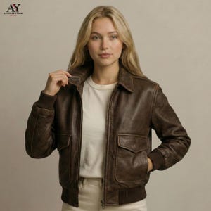 May include: A brown leather bomber jacket with a zip-up front, two front pockets, and ribbed cuffs and hem. The jacket has a classic collar and is worn over a white t-shirt. The jacket is a stylish outerwear piece.