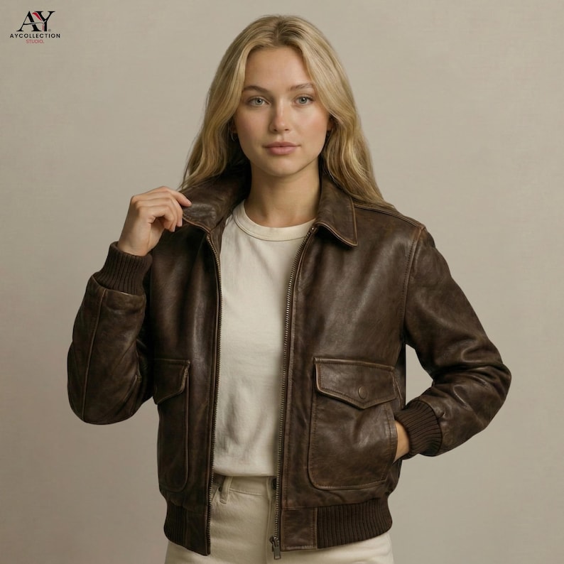 May include: A brown leather bomber jacket with a zip-up front, two front pockets, and ribbed cuffs and hem. The jacket has a classic collar and is worn over a white t-shirt. The jacket is a stylish outerwear piece.