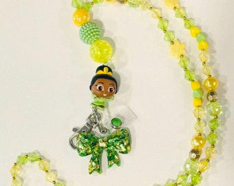 May include: A beaded lanyard in shades of yellow and green, featuring a cartoon character charm and a glittery green bow. The lanyard is adorned with various bead shapes, including round and faceted beads.
