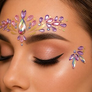 May include: Close-up of a person's face with closed eyes, adorned with face jewels. The jewels are in shades of purple, pink, and iridescent, arranged in a floral design above the eyebrow and on the cheek.