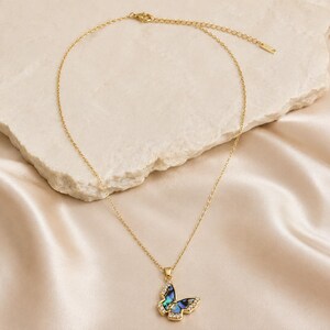 May include: A gold-colored necklace with a butterfly pendant. The butterfly pendant has a blue and green iridescent center with clear crystal accents. The necklace is displayed on a cream-colored stone and satin fabric.