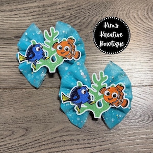 May include: Two turquoise hair bows with glitter accents. Each bow features a cartoon image of Dory and Nemo from the movie "Finding Nemo" on a green coral-shaped background. The bows are on a wooden surface. The text "Kim's Kreative Boutiqe" is in a black circle.