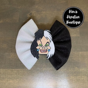May include: A handmade hair bow featuring a Cruella de Vil character design. The bow is split into two sections, with one side white and the other black. The Cruella graphic is centered on the bow, and the text "Kim's Kreative Bowtique" is in a circle.