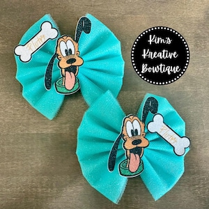 May include: Two turquoise blue tulle bows with a glitter finish. Each bow features a cartoon image of Pluto, a dog from Disney's Mickey Mouse, with a bone that says "Pluto".