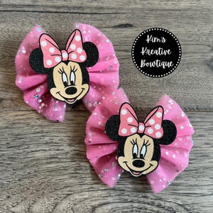 May include: Two pink hair bows with glitter accents and Minnie Mouse faces. Each bow features a pink polka dot bow and black mouse ears. The bows are set against a wooden background. The text "Kim's Kreative Boutique" is in a black circle.