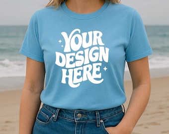 May include: A light blue t-shirt with the text "YOUR DESIGN HERE" in white, displayed in a retro font. The model is wearing blue jeans. The background shows a beach and ocean.