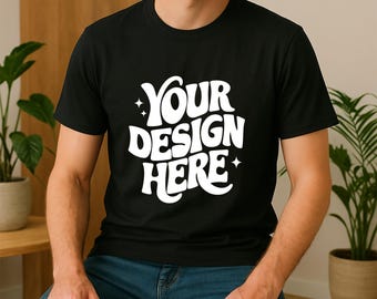 May include: Black crew neck t-shirt with the text "YOUR DESIGN HERE" in white, a mock-up for custom designs. The shirt is worn with blue jeans. The image is taken indoors with plants in the background.