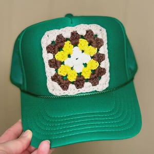 May include: Green trucker hat with a brown, white, and yellow crocheted square patch.