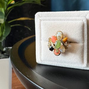 May include: A gold-toned ring featuring a cluster of multi-colored oval cabochon gemstones, including coral, green jade, white opal, and tiger's eye, set in a decorative metal frame with intricate filigree details and small clear accent stones.