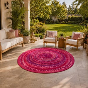 May include: A round, braided area rug featuring a vibrant mix of pink, magenta, and purple tones. The rug has a circular, concentric pattern created by tightly woven fabric strands, providing a textured surface that sits flat on the floor.
