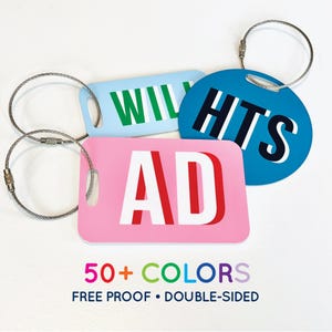 May include: A collection of luggage tags in various colors, including light blue, pink, and blue. Each tag features bold white lettering with red accents, spelling out "WIL", "AD", and "HITS". The tags are attached to silver metal loops. The text "50+ COLORS" and "FREE PROOF • DOUBLE-SIDED" is also visible.