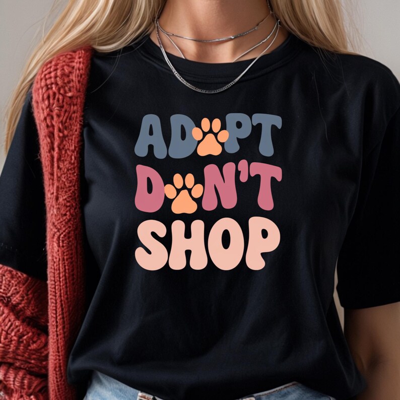 May include: Black t-shirt with the text "ADOPT DON'T SHOP" in colorful retro font with paw prints.