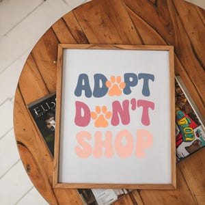 May include: A framed print with the text "Adopt Don't Shop" in a retro font style. The words are in shades of blue, pink, and orange with paw prints in between each word.