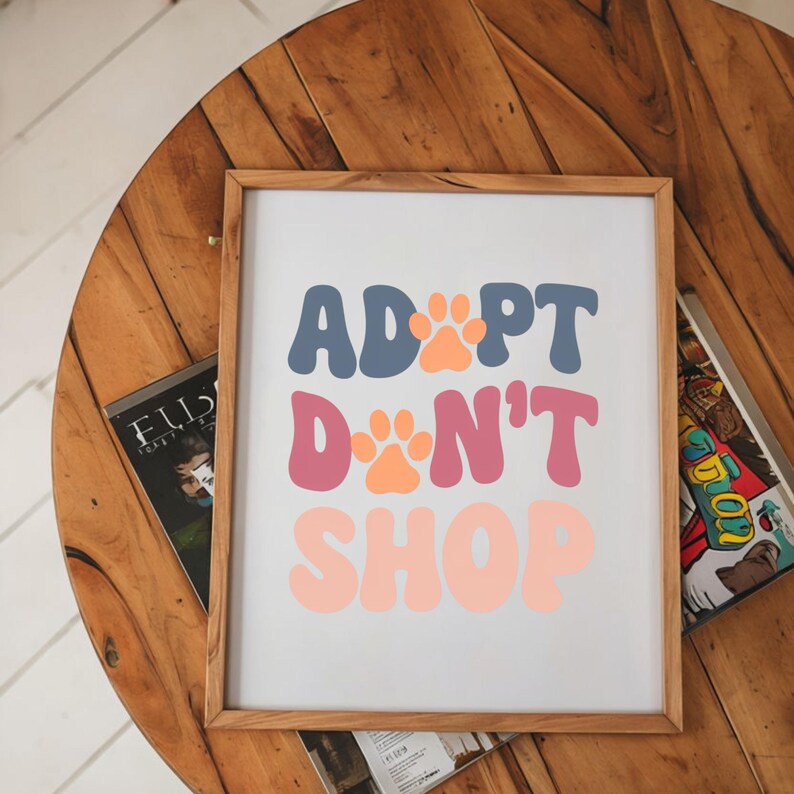 May include: A framed print with the text "Adopt Don't Shop" in a retro font style. The words are in shades of blue, pink, and orange with paw prints in between each word.