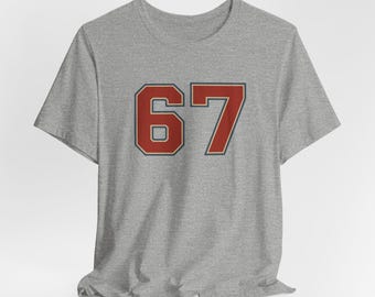 May include: A heather gray t-shirt featuring the number "67" in a bold, red font with a dark red outline. The shirt has short sleeves and a crew neck. The fabric appears soft and slightly wrinkled.