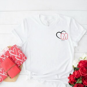 Puede incluir: Una camiseta blanca con un diseño de corazón negro y rojo. El diseño incluye las letras "CM" y la fecha "MR 29-21". La camiseta se muestra sobre una superficie de madera blanca, con artículos con estampado de corazones rojos y rosas rojas.