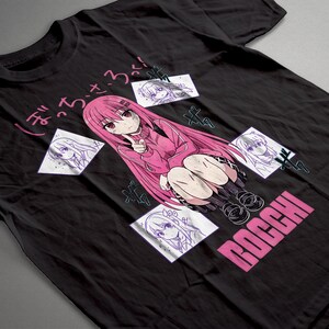 May include: A black short-sleeve t-shirt featuring a central graphic of a pink-haired anime character in a pink sweater. The design includes three smaller white square sketches of the character, Japanese text, and the word BOCCHI in pink letters.