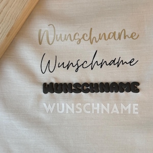 Könnte beinhalten: Nahaufnahme eines hellen Stoffes mit dem Wort "Wunschname" in vier verschiedenen Stilen: Gold, schwarze Schrift, dunkelgrau 3D und Weiß. Der Text ist wahrscheinlich für personalisierte Artikel.