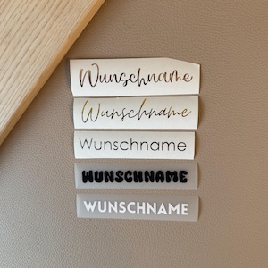Könnte beinhalten: Fünf rechteckige Aufkleber, die vertikal angeordnet sind und jeweils das Wort "Wunschname" in verschiedenen Schriftarten und Farben zeigen. Die Aufkleber umfassen metallisch goldene Schreibschrift, goldene Kursivschrift, schwarze Serifenschrift, fette schwarze Blasenbuchstaben und weiße serifenlose Schrift.