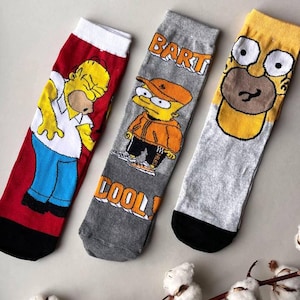 Puede incluir: Tres pares de calcetines coloridos con personajes de la caricatura Los Simpson. Los calcetines son rojos, grises y amarillos con detalles negros. Los personajes en los calcetines incluyen a Homero, Bart y un personaje amarillo con la cara marrón.