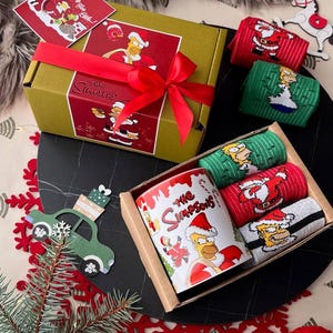 May include: A festive gift set with The Simpsons theme, including a mug, socks, and a box tied with a red ribbon. The socks are in red, green, and grey. The mug and box are decorated with the show's logo and Christmas imagery.